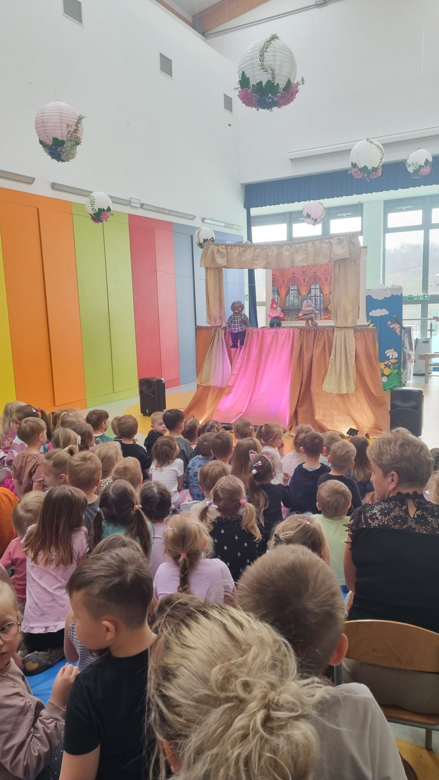 Dzieci siedzą na holu przedszkola i oglądają kukiełkowe przedstawienie na scenie z beżowymi kurtynami. Na scenie widać kilka kukiełek oraz podświetloną na różowo dekorację. Przestrzeń jest jasna i kolorowa, a z sufitu zwisają ozdoby.