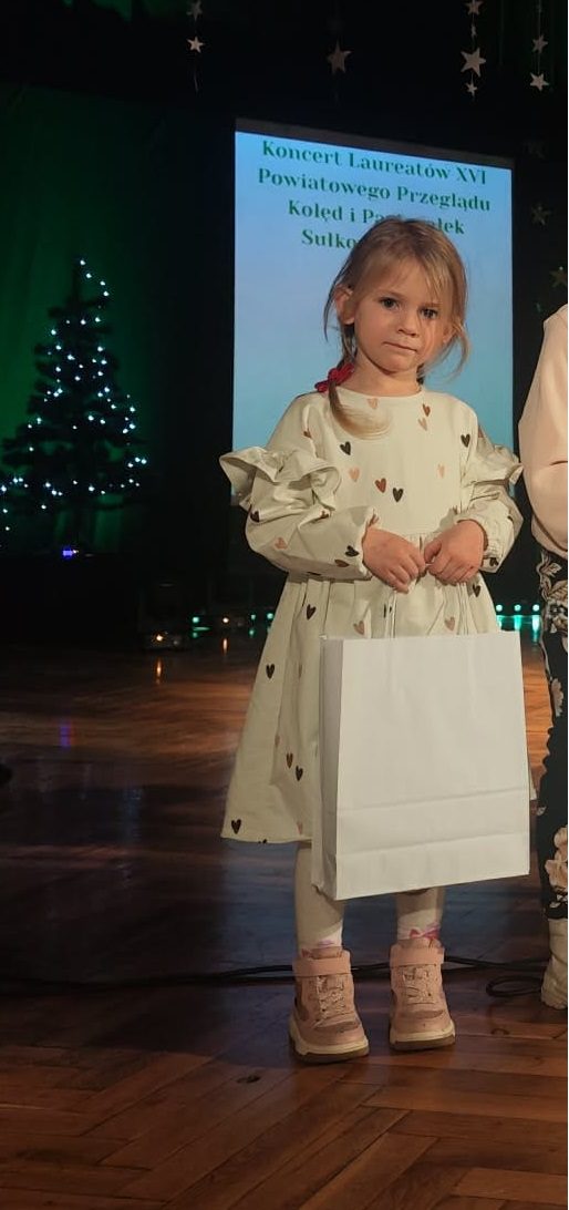 Dziewczynka stoi na scenie podczas koncertu laureatów XVI Powiatowego Przeglądu Kolęd i Pastorałek w Sułkowicach. Trzyma pamiątkową torbę z nagrodą, w tle świąteczna dekoracja i ekran z nazwą wydarzenia.